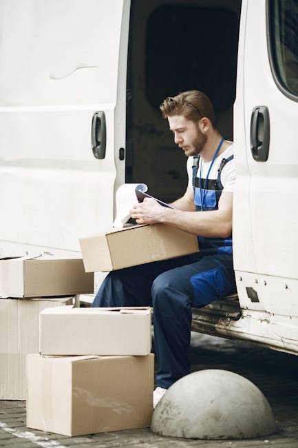 A man with a beard and wearing a black cap, white polo shirt, and jeans is kneeling on the pavement next to an open cardboard moving box inside a van. He is using a black marker to label the box, whic