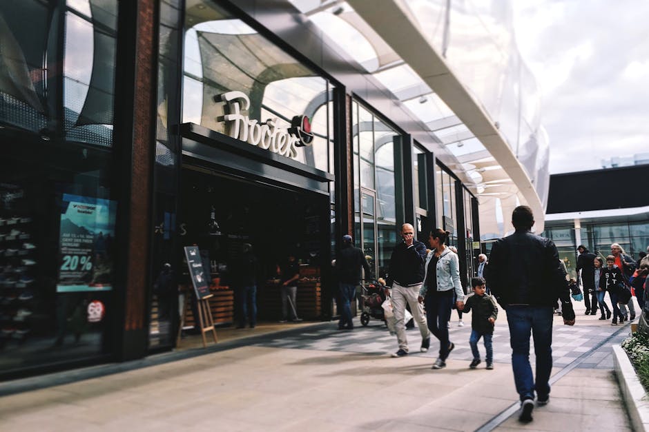 The image shows the exterior of a modern shopping centre with large glass windows and a curved architectural structure under a cloudy sky. Multiple pedestrians are walking along the paved pathway outside, including a man and woman engaged in conversation, a young child holding hands with an adult, and others passing by, some with shopping bags. Inside the shopping centre, partially visible through the glass, are signs of retail spaces such as a Foodies store, with a wooden display stand positioned near the entrance. The scene captures a typical busy day with shoppers, and the area appears well-lit with natural daylight. This setting relates to house removals and furniture transport, reflecting the logistical process of relocating household items through professional services like those offered by Man and Van Brent Cross, which handle packing and moving logistics for residents in the Brent Cross area.