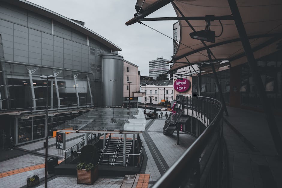 A view of the exterior of a modern shopping centre with a curved balcony on the right side, partly covered by a canopy. The balcony has a metal railing and is adjacent to a commercial building with large glass windows and metal cladding. Below, the open walkway features steps leading down to street level, with a few people walking in the distance. Potted plants are placed near the escalator, which is visible in the centre of the image. An electronic sign for a food venue named 'hollywood + bowl' is mounted on the building, and there is a visible pedestrian crossing with traffic lights in the background. The scene is captured in daylight with overcast skies, indicating a typical day of activity at the shopping complex, which might be relevant for home relocation or furniture transport planning by Man and Van Brent Cross.