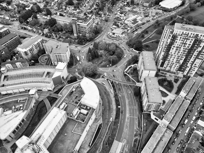 An aerial black-and-white photograph showing a busy urban area with multiple multi-storey buildings, roads, and traffic. The image captures an intricate network of streets, including a roundabout at the intersection of several roads, with vehicles visible moving along the roads and parked along the edges. In the foreground, a large commercial or institutional building with flat roofs and multiple loading bays is visible, with some trucks and vans parked nearby, indicative of loading or unloading activities. Adjacent to this building, there are smaller structures, possibly offices or service units, with materials such as cardboard boxes, plastic wrapping, and moving blankets visibly stacked on the pavement, suggesting active packing and home relocation preparations. The scene also features a section of pavement and a sidewalk where some objects are being carried or moved, in alignment with house removals and packing processes. The overall environment is well-lit, with natural light casting shadows from the buildings and trees, illustrating the logistics involved in furniture transport and moving services. Man and Van Brent Cross undertakes such loading, packing, and transport operations in these areas, supporting efficient removals near Brent Cross.