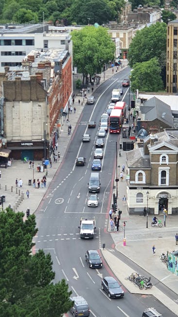 An aerial view of a busy residential street in Hendon Way near Brent Cross, showing multiple cars queuing in a single lane, with some vehicles moving slowly or waiting at traffic signals. On the left side of the road, a sidewalk is populated with pedestrians walking, including a person pushing a stroller and others carrying shopping bags. Adjacent to the sidewalk, a row of red-brick commercial and residential buildings is visible, with shopfronts and upper-floor windows. On the right side, a small, light-colored house with a pitched roof and decorative trim is situated at the corner, near a bicycle rack with several bikes parked. The street is lined with mature trees providing shade and greenery, creating a pleasant urban environment. The overall scene depicts typical street traffic and pedestrian activity, consistent with an area where local removals services might operate, supporting professional house and furniture transport, packing, and loading processes.