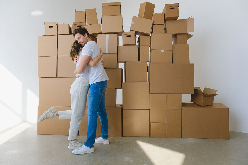 A man with dark hair and a light beard wearing a white t-shirt is surrounded by numerous cardboard boxes of various sizes, some stacked on top of each other and others scattered around, in an indoor s